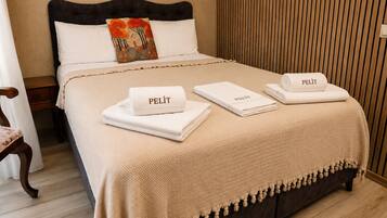 Una cama bien tendida con cabecera oscura, almohadas blancas y una almohada decorativa con una escena de naturaleza. Hay tres toallas dobladas con la palabra 'PELIT' y una manta beige texturizada con flecos al pie de la cama.