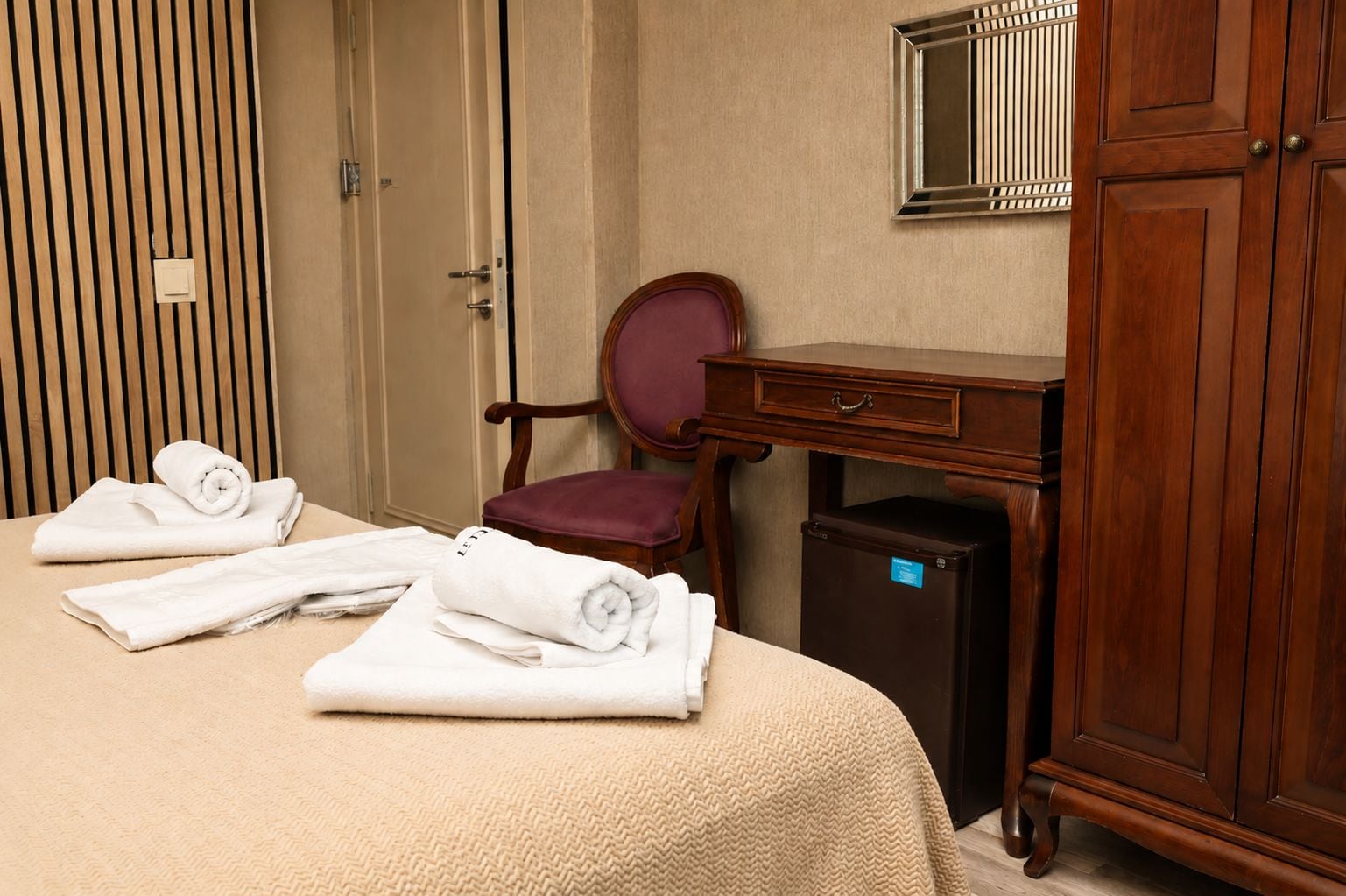 A hotel room with a bed, a desk, a chair, a wardrobe, and a small refrigerator.