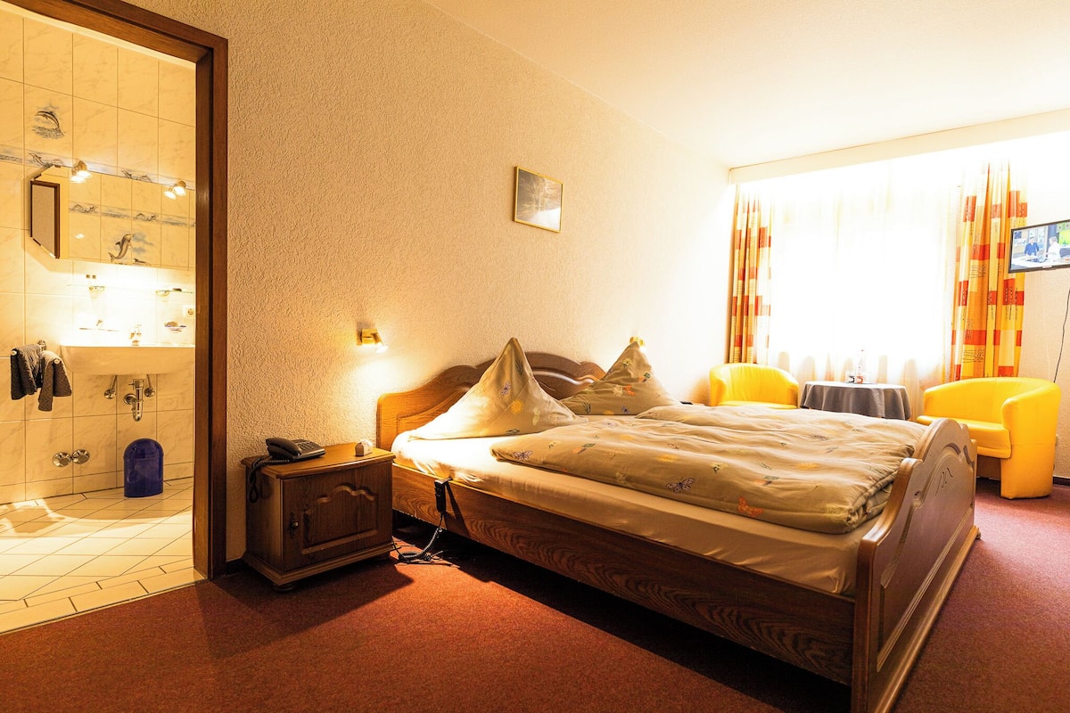 A hotel room with a wooden bed, a bedside table with a lamp, a small table with a TV, and a bathroom with a mirror and sink.