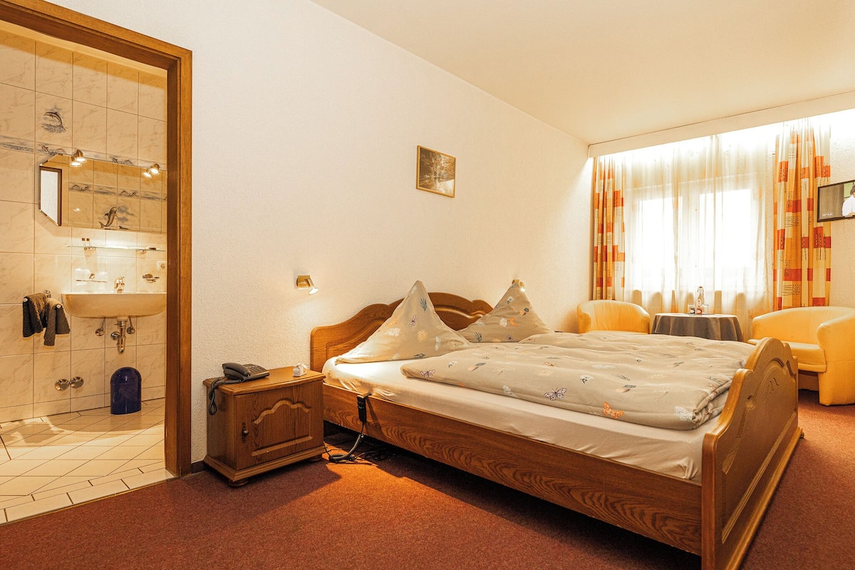 A hotel room with a wooden bed, a bedside table with a phone, a small cabinet, a bathroom with a sink and mirror, and a window with curtains.