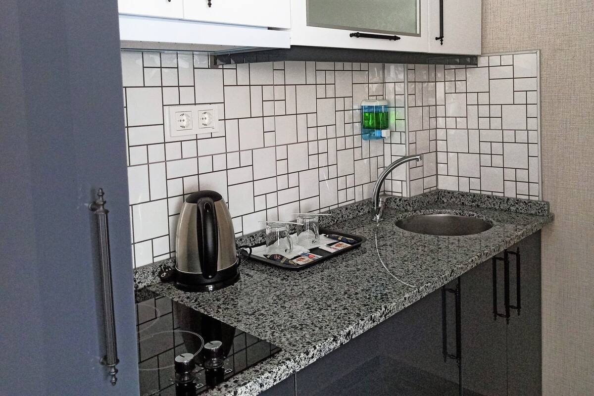 A compact kitchen with a granite countertop, a stainless steel kettle, a sink, and a tiled backsplash.