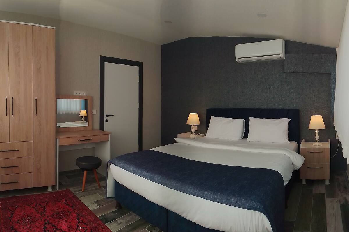 A hotel room with a bed, bedside tables, a desk, a wardrobe, and an air conditioning unit.