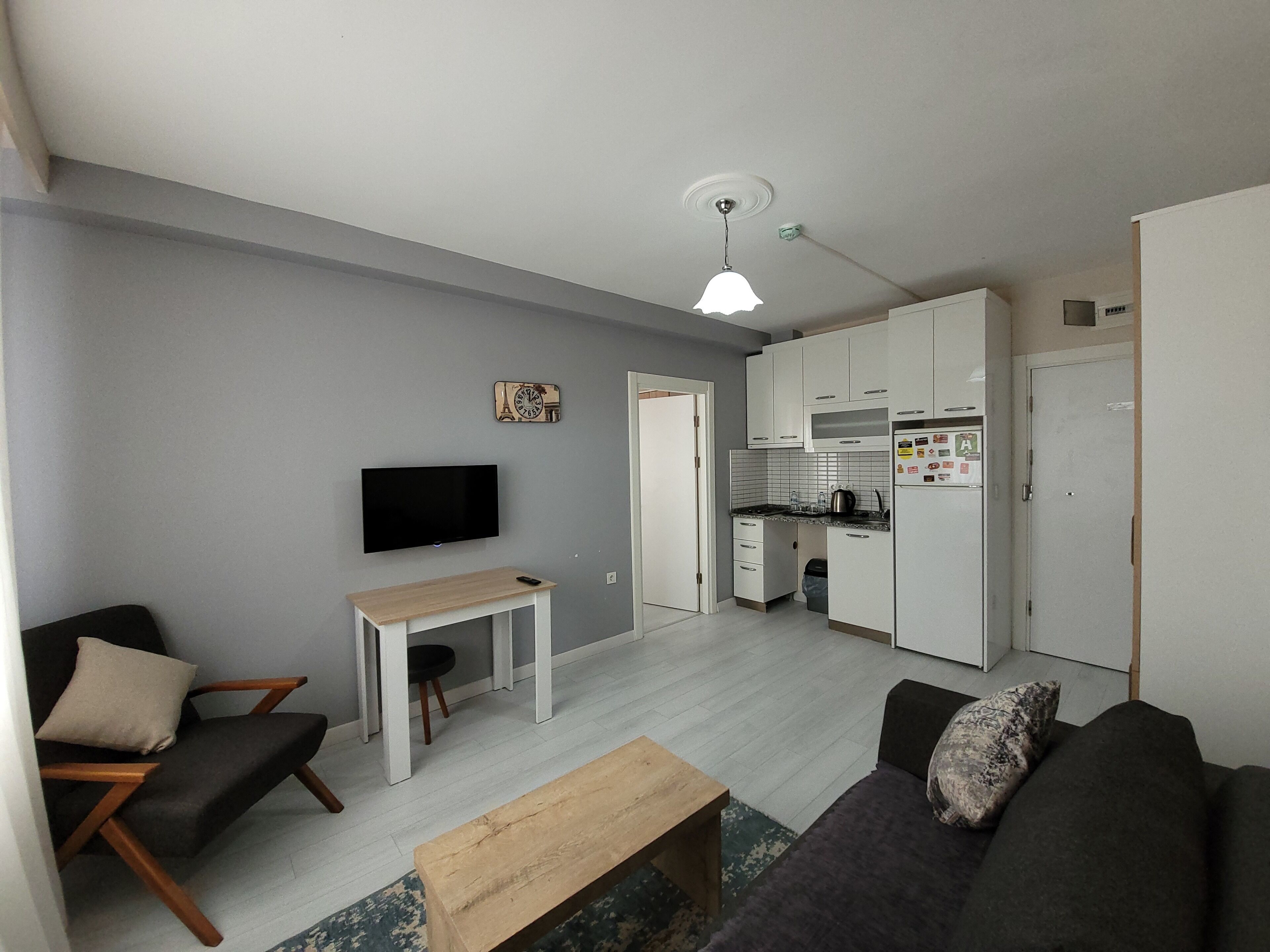 Una sala de estar moderna con televisor, sofá y mesa de centro. Se ve una cocina al fondo con gabinetes y electrodomésticos blancos.