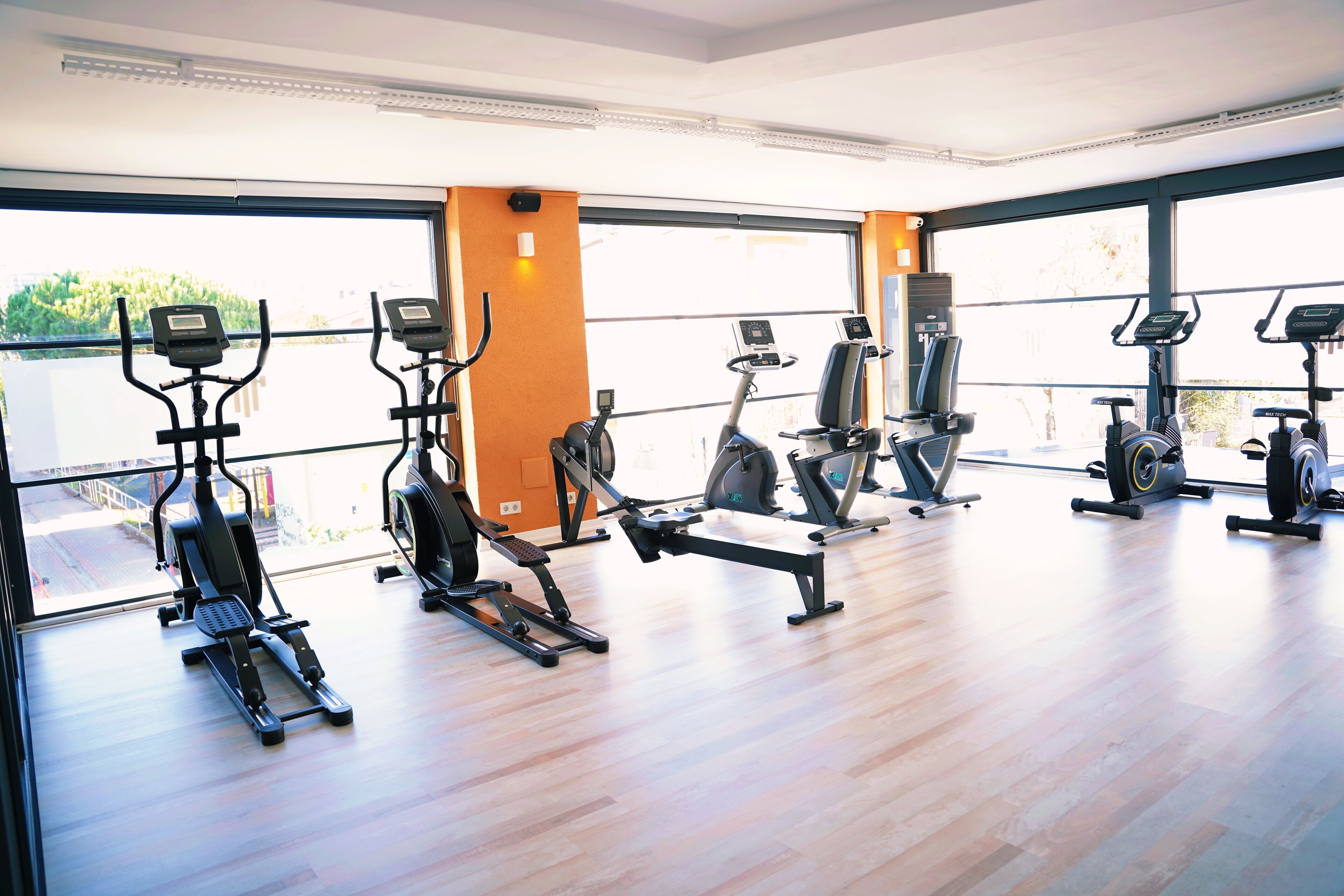 A gym with various exercise machines, large windows, and a wooden floor.