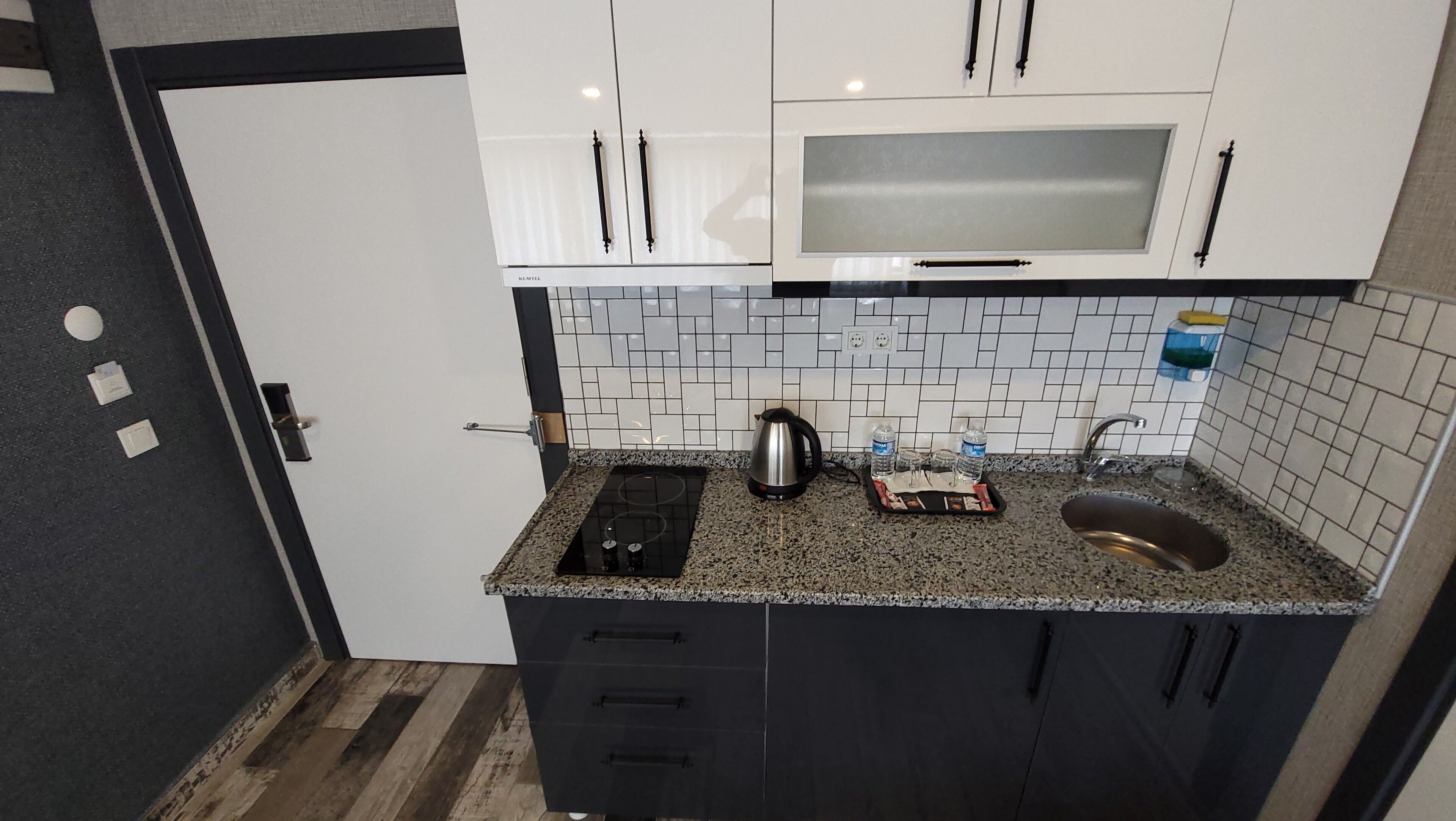 A compact kitchen with white cabinets, a black countertop, and a microwave. There are three water bottles on the counter.