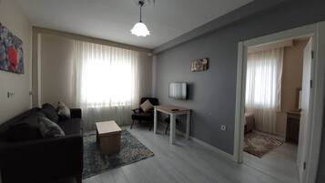 Una sala de estar con un sofá oscuro, una mesa de centro de madera, un televisor sobre un mueble y una puerta que da a otra habitación.
