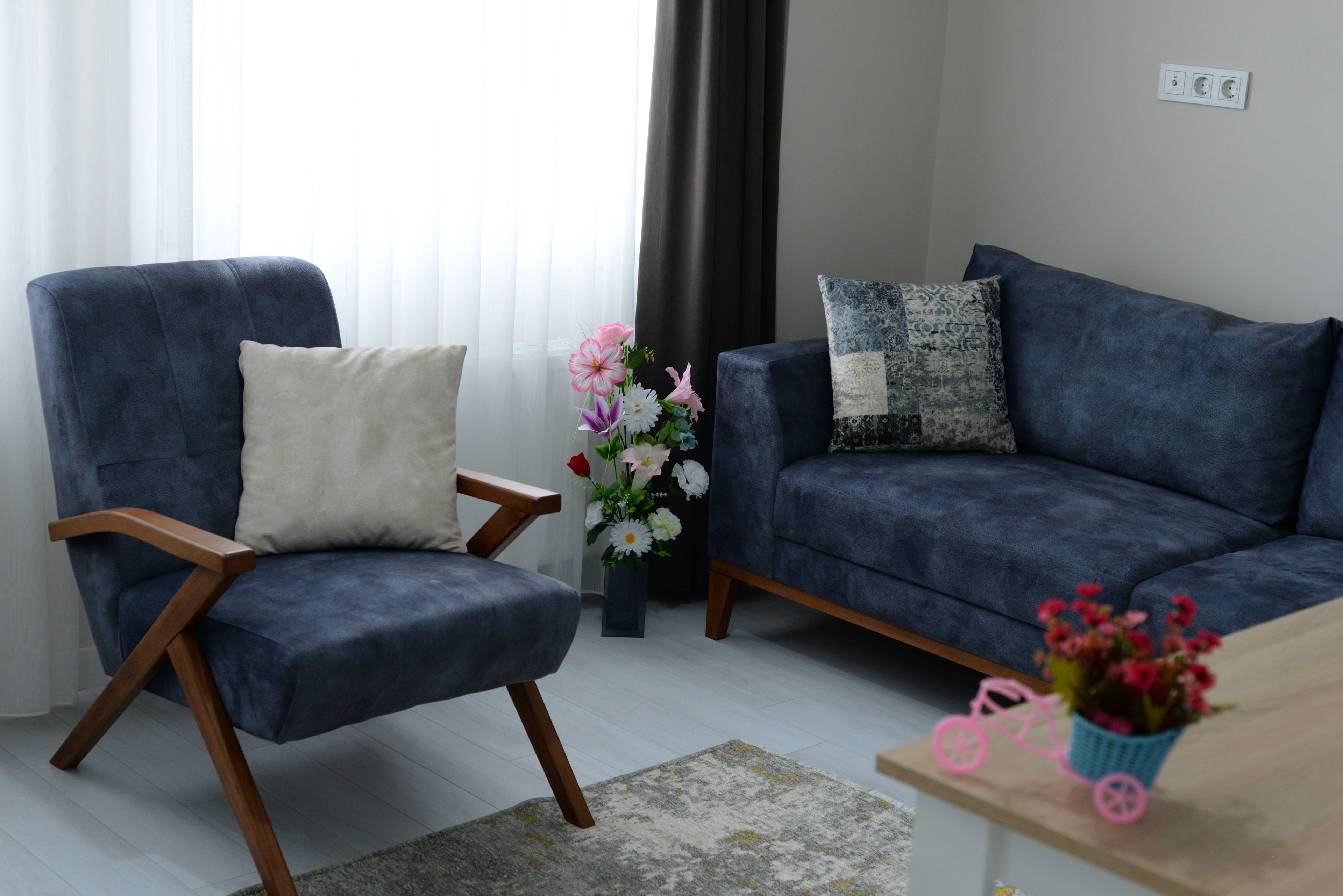 A living room with a blue armchair, a blue sofa, a small table with a pink toy and flowers, and a vase with flowers.