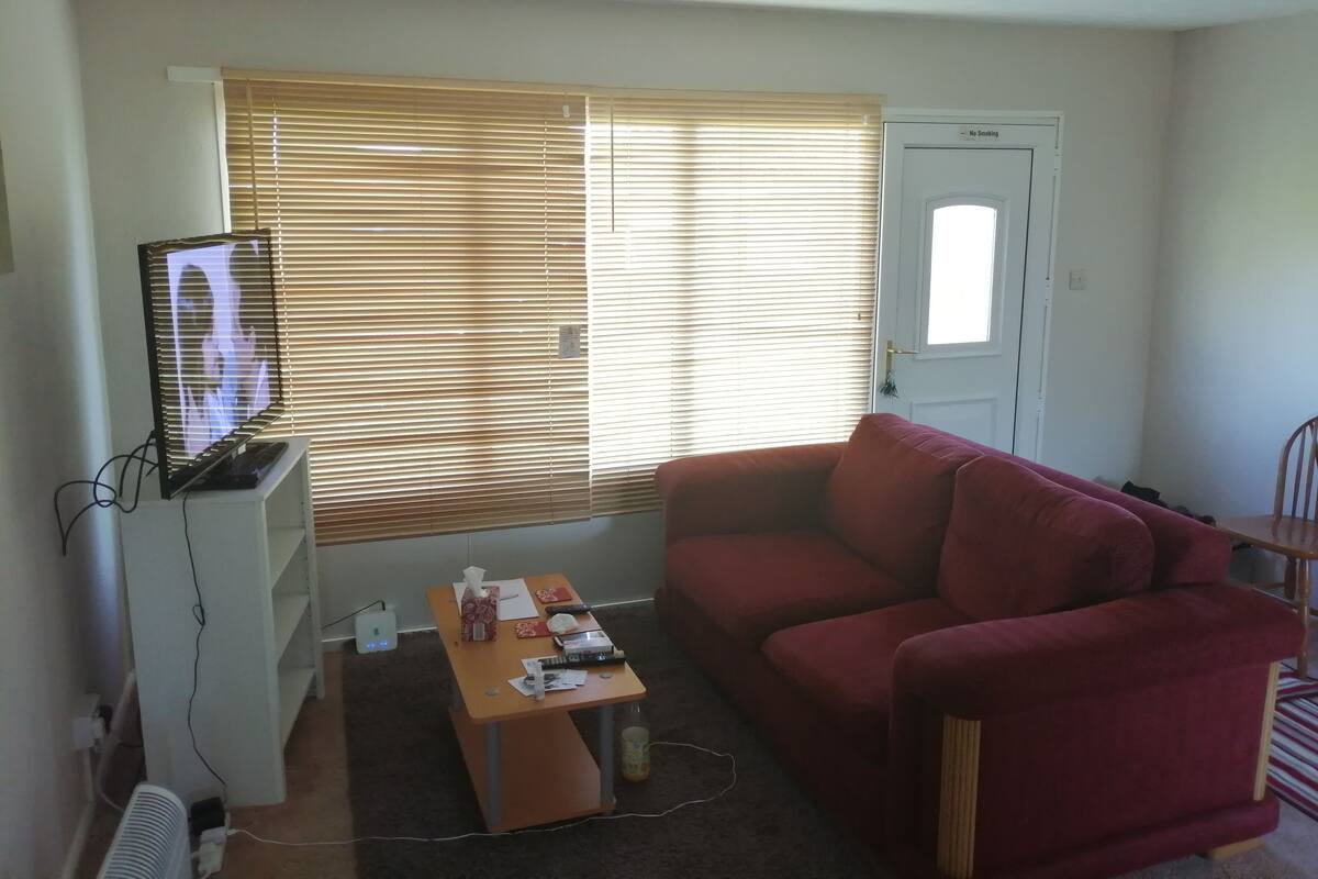 A living room with a red sofa, a TV on a stand, a small table with items, and a window with blinds.