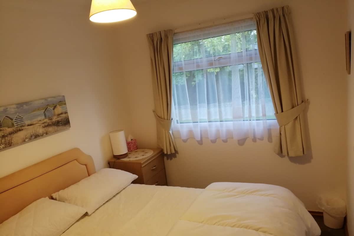 A bedroom with a bed, a nightstand with a lamp, a painting on the wall, and a window with curtains.