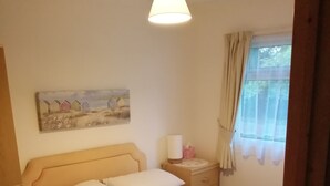 Uma cama bem arrumada com lençóis brancos, uma mesa de cabeceira com abajur, um quadro na parede e uma janela com cortinas.