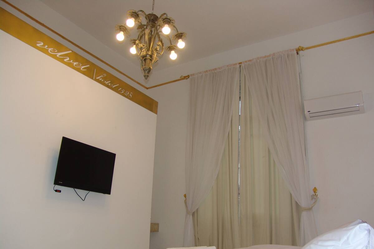 Una habitación de hotel con cama, televisión montada en la pared, candelabro y cortina.