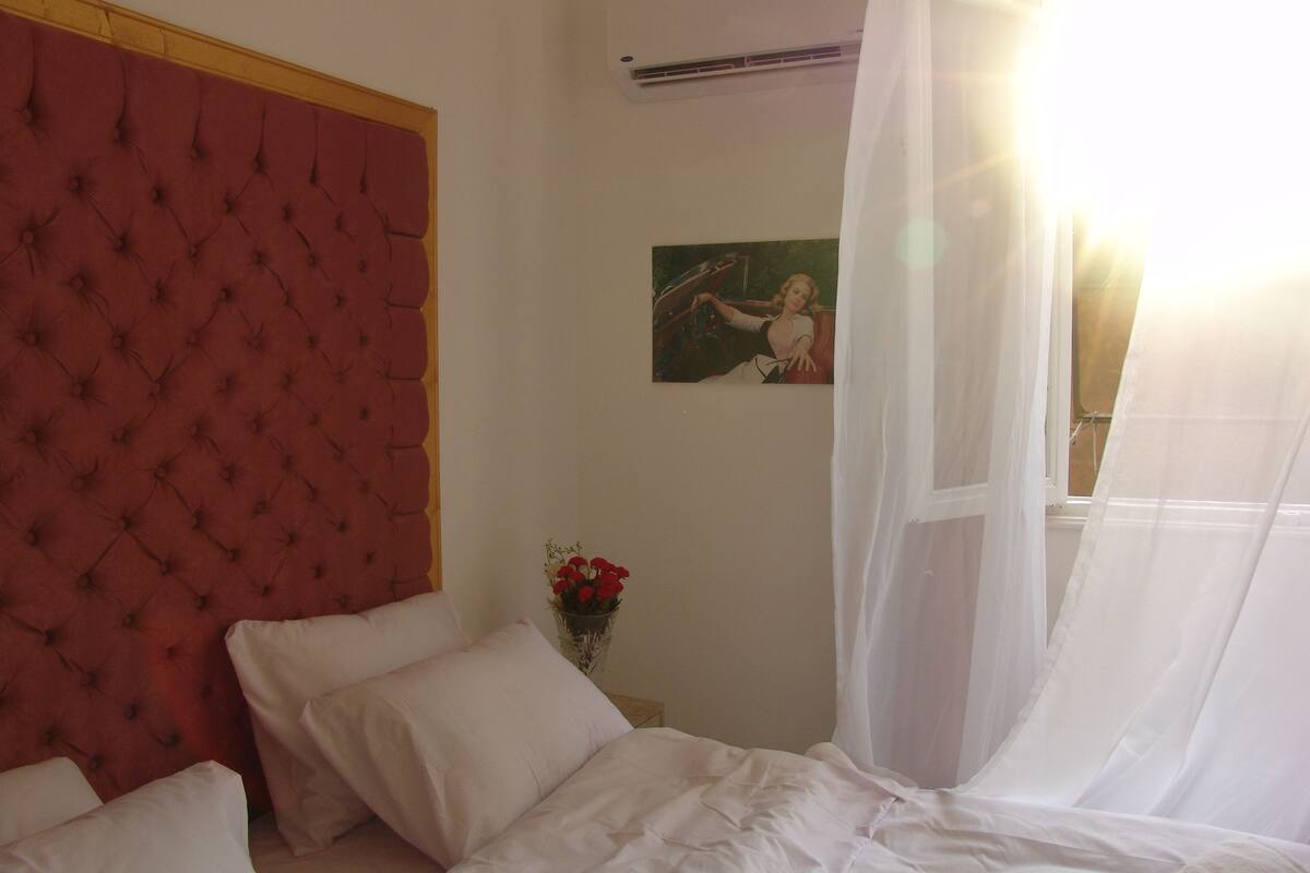 Un dormitorio con cabecera capitonada, cama con ropa de cama blanca, una mesita de noche con un jarrón de flores rojas y un cuadro en la pared.