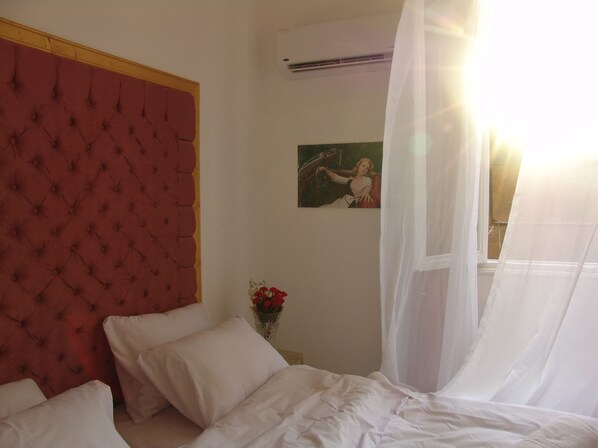 Un dormitorio con cabecera capitonada, cama con ropa de cama blanca, una mesita de noche con un jarrón de flores rojas y un cuadro en la pared.