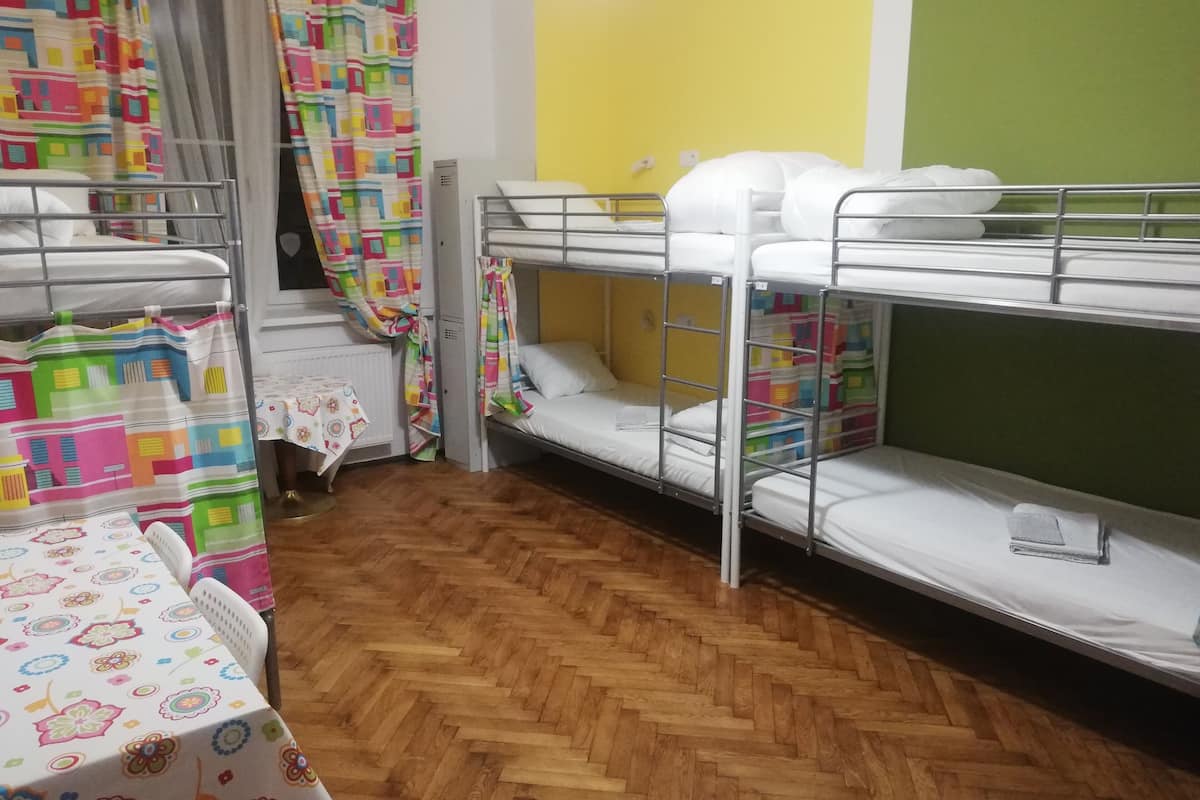 A dormitory room with bunk beds, a wooden herringbone floor, a table with a floral tablecloth, and colorful curtains.