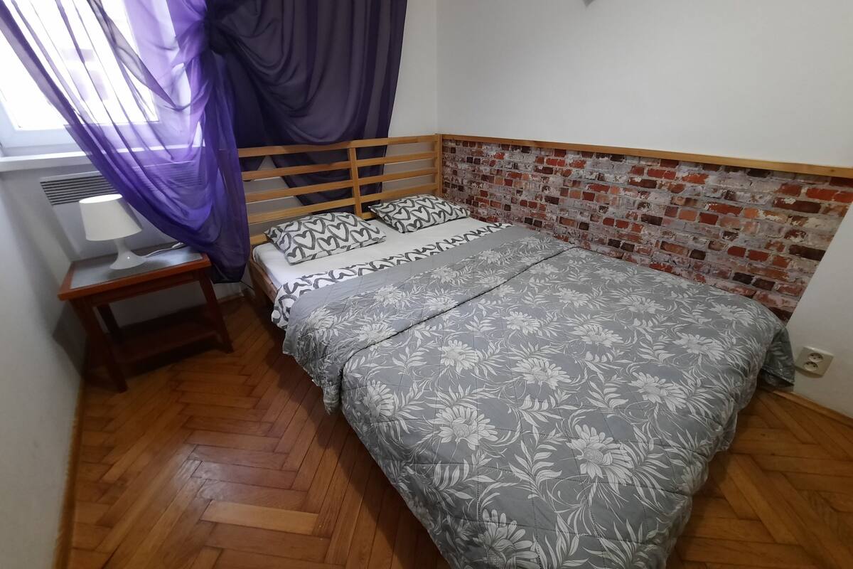 A bedroom with a wooden bed frame, floral bedding, a nightstand with a lamp, and a brick-patterned wall.