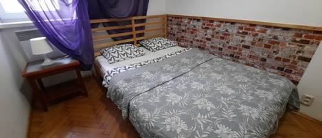 Un dormitorio con estructura de cama de madera, ropa de cama floral, una mesita de noche con lámpara y una pared con diseño de ladrillo.