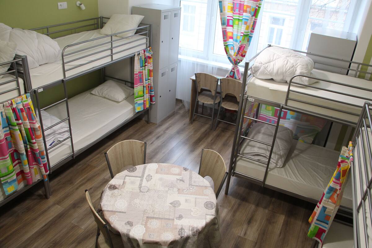 A dormitory room with bunk beds, a round table, and a window with curtains.