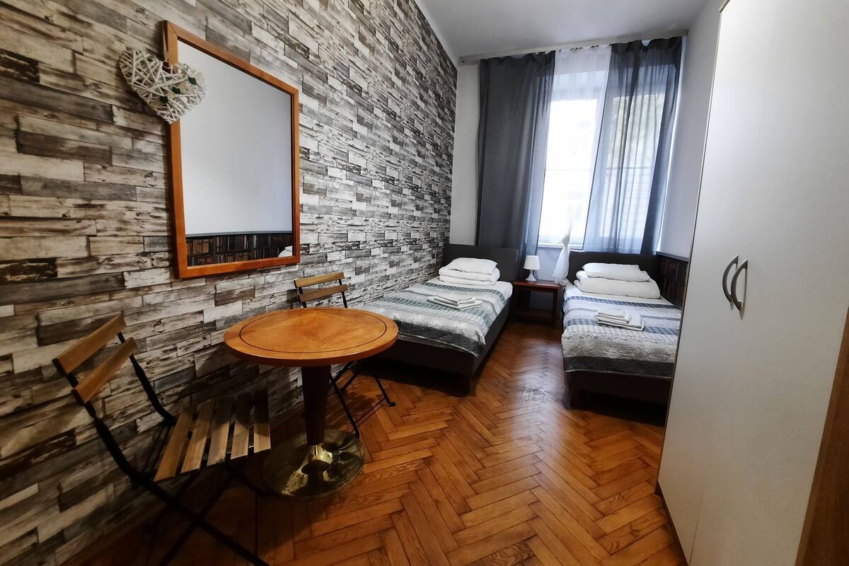 A room with a herringbone wood floor, a round wooden table, two single beds with white linens, a wooden bench, a mirror on the wall, and a window with curtains.