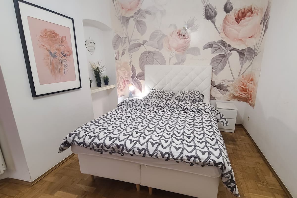 A bedroom with a bed, a bedside table, a nightstand, a framed picture, and a floral wallpaper.