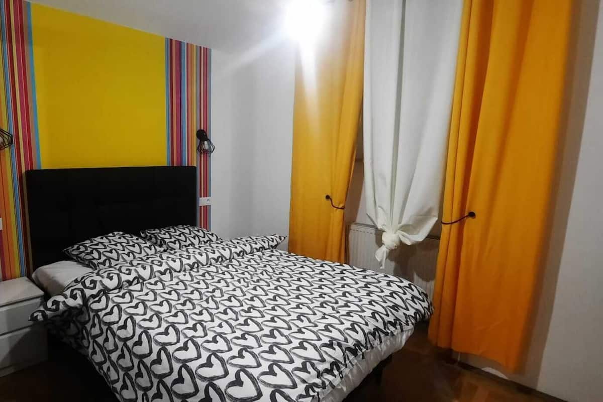 A bedroom with a bed, a nightstand, a wall with a colorful striped pattern, and a door with curtains.