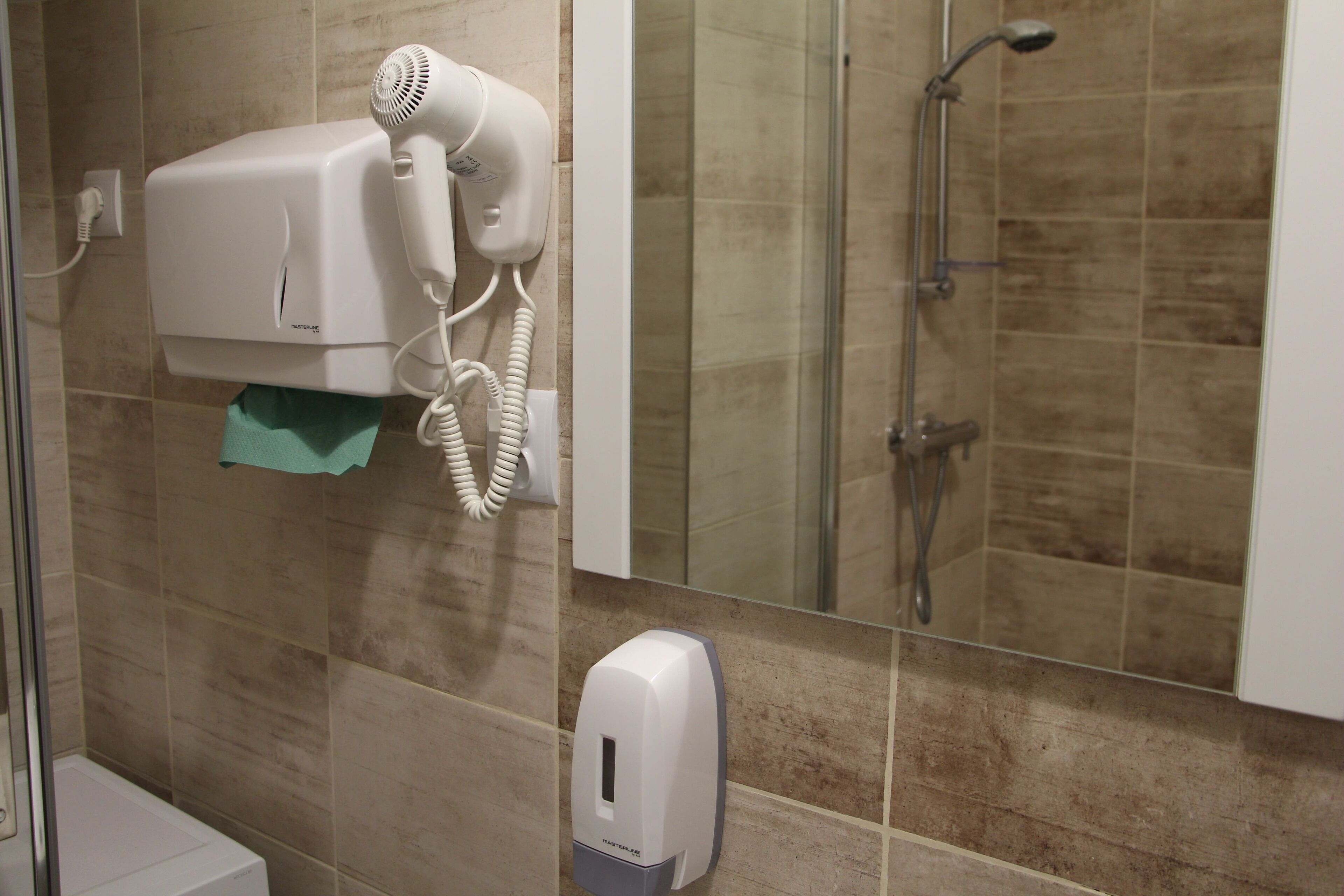 A bathroom with a hairdryer mounted on the wall, a towel dispenser, and a shower area with a glass door.