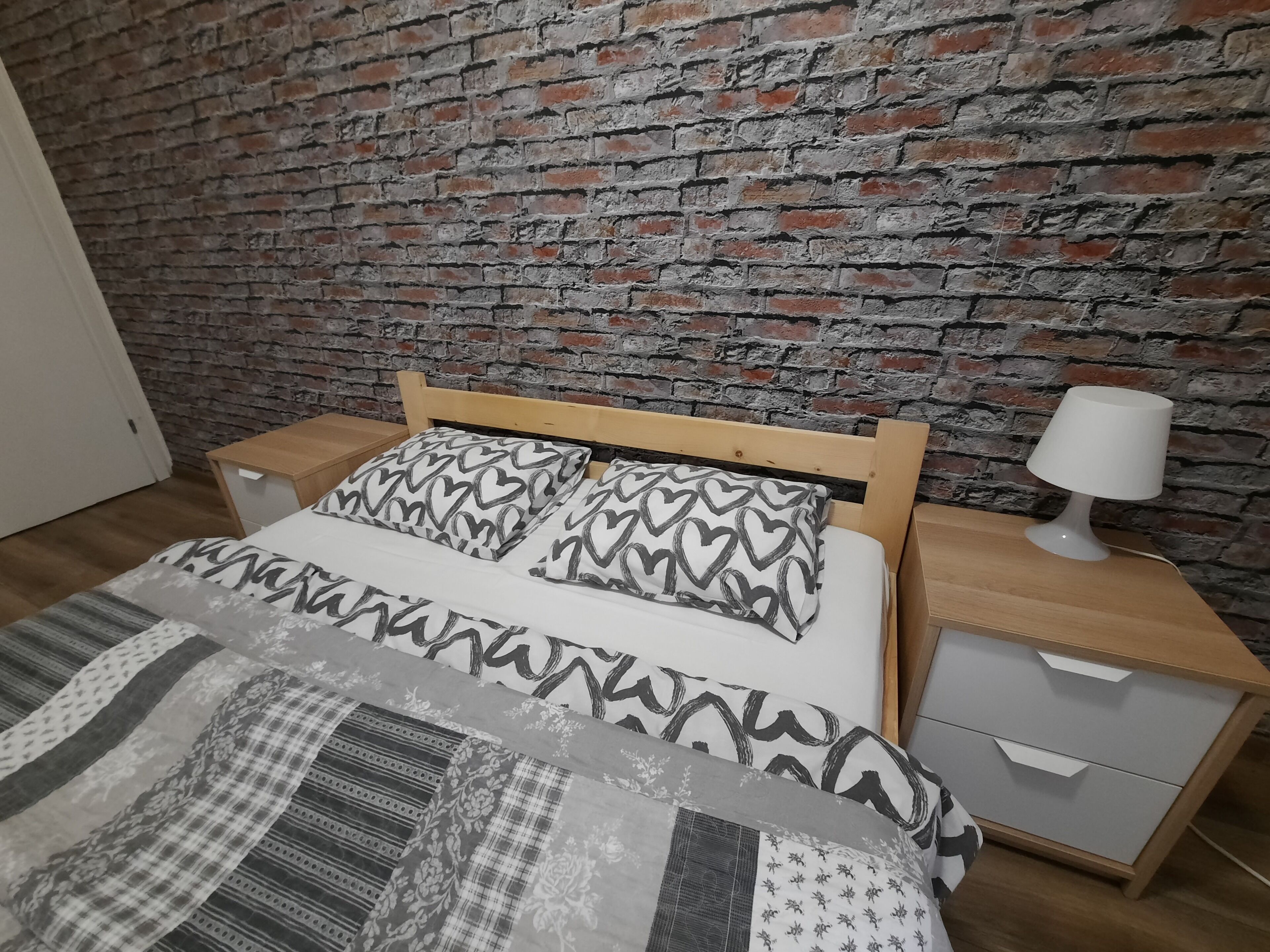 Ein Bett mit gemusterten Betttextilien, ein hölzernes Kopfteil, ein Nachttisch mit Lampe und ein Beistelltisch mit Schublade.