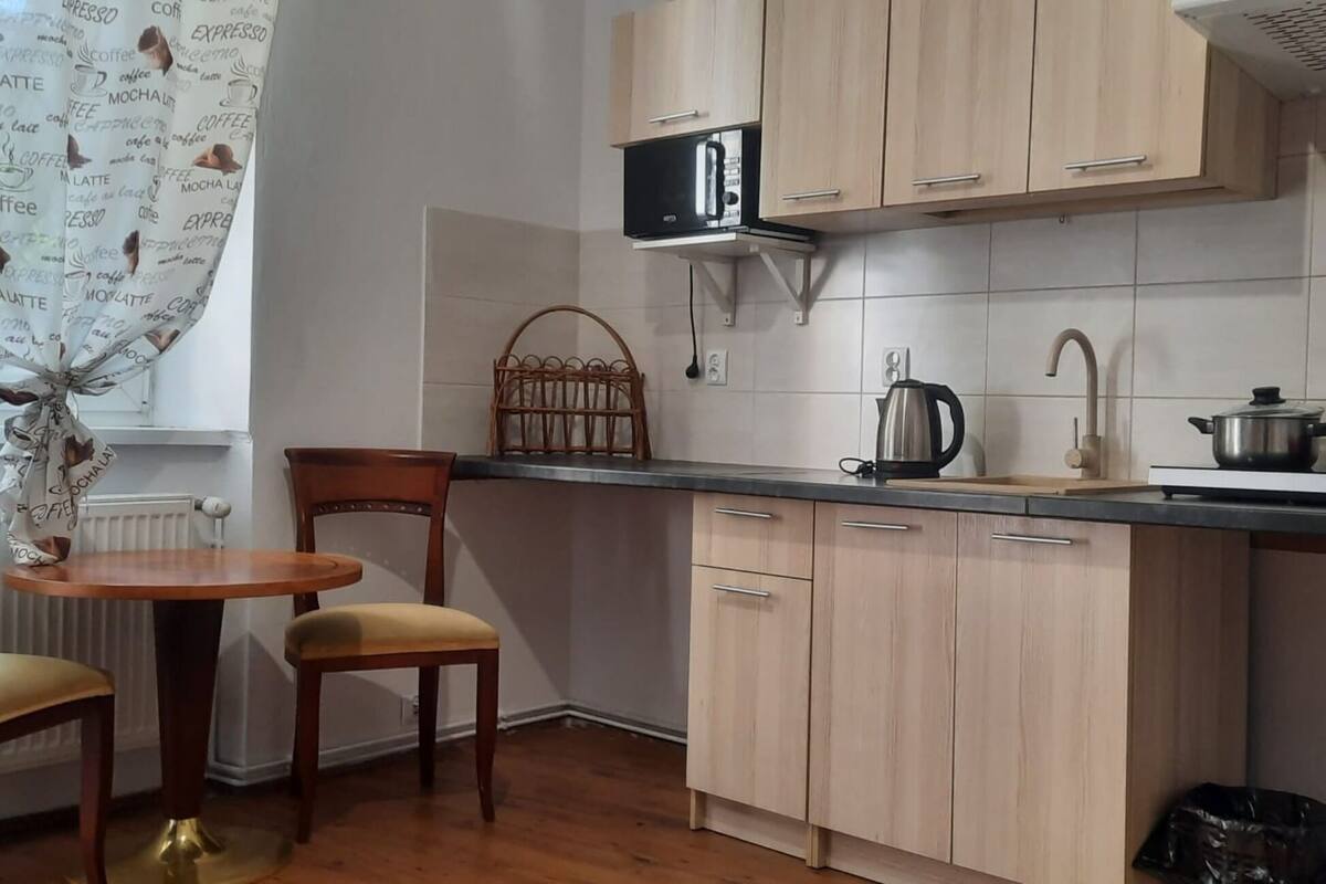 A small kitchen with wooden cabinets, a microwave, a kettle, and a sink. There is a wicker chair and a small table with a chair.