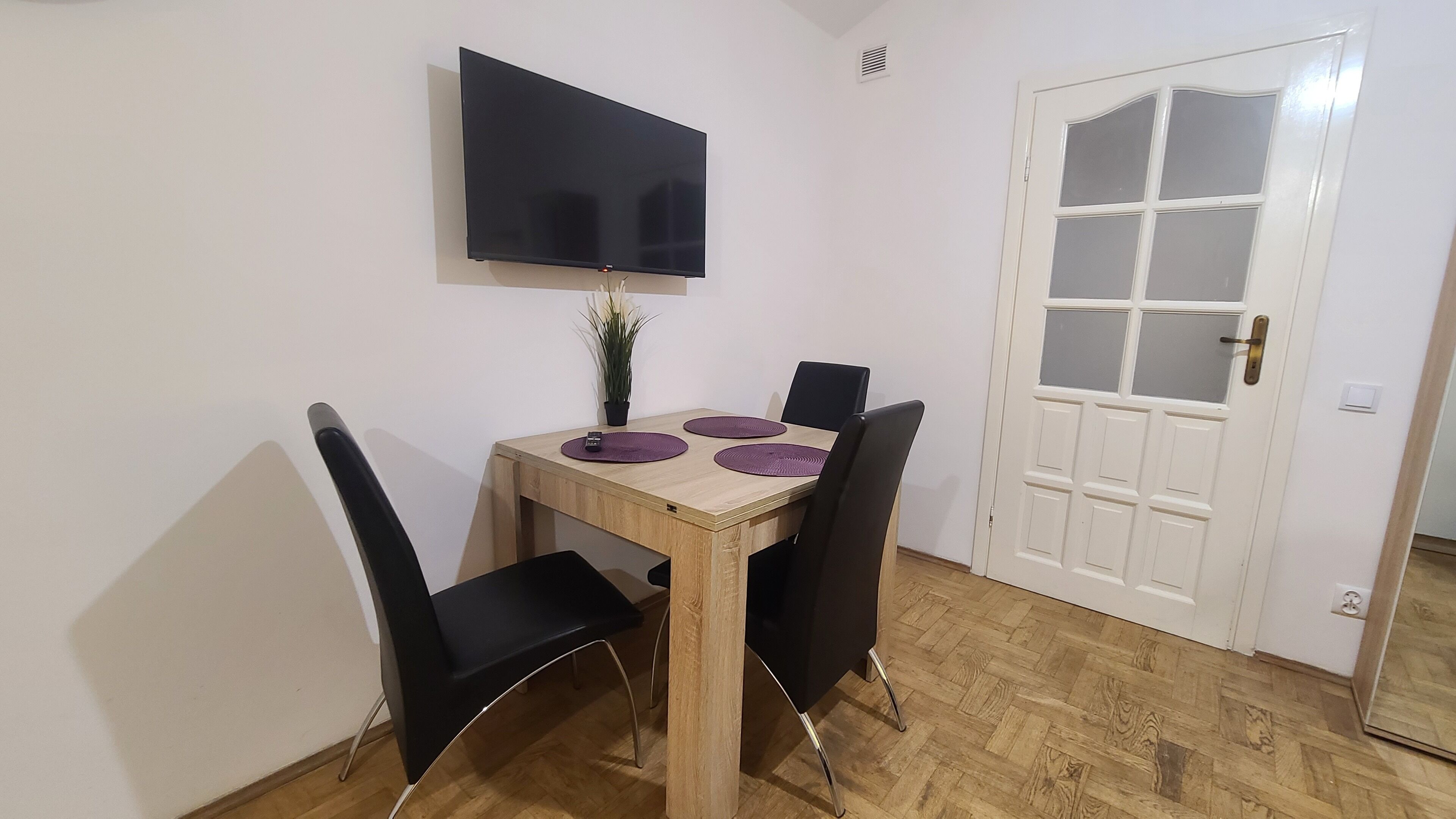 A dining area with a wooden table, black chairs, a flat-screen TV mounted on the wall, and a white door with glass panels.