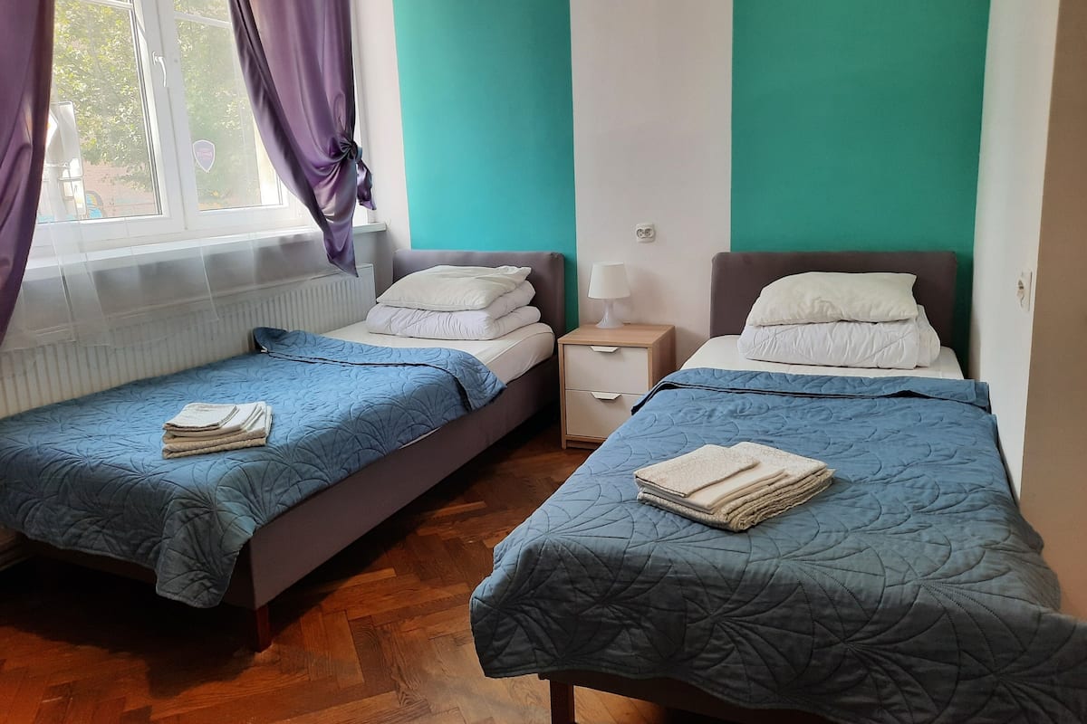 A room with two single beds, each with a blue quilt, a bedside table with a lamp, and a window with curtains.