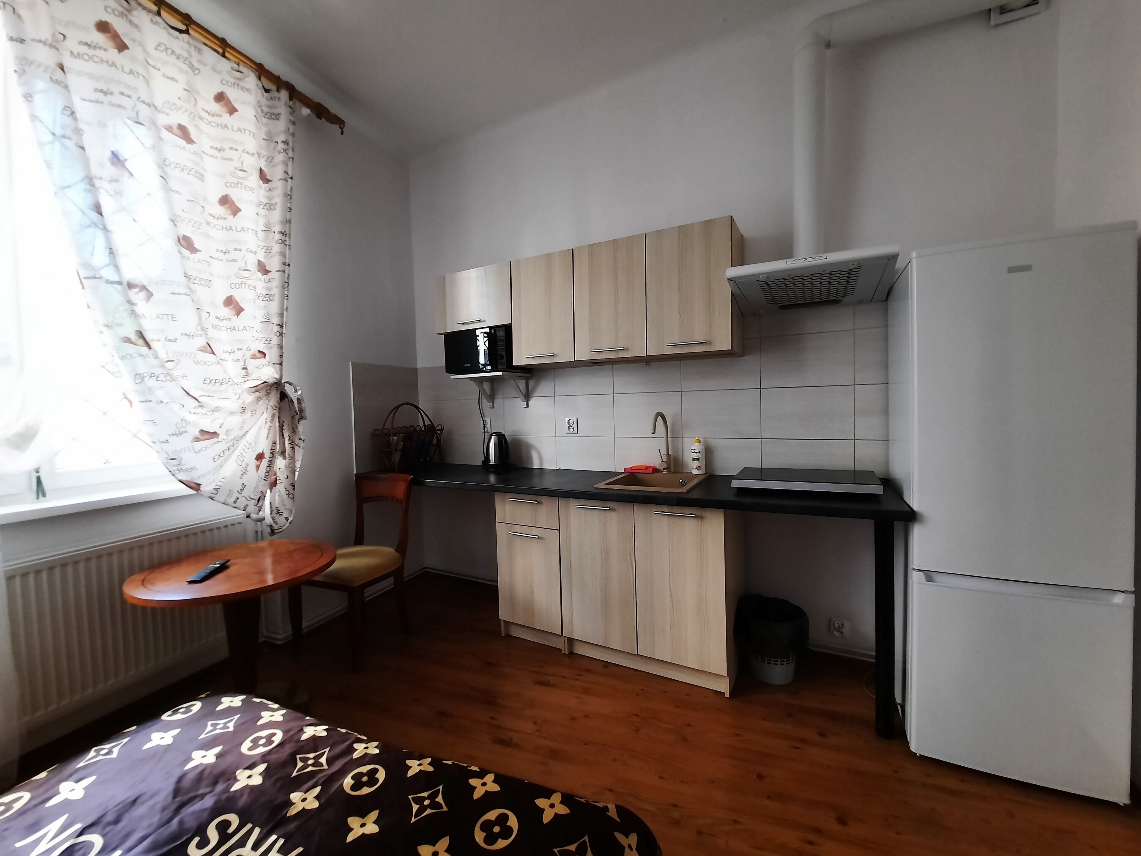 Eine kompakte Küche mit Mikrowelle, Spüle und Kühlschrank. Ein kleiner Esstisch und ein Stuhl bieten Platz zum Essen. Der Raum verfügt über Holzböden und helle Wände.
