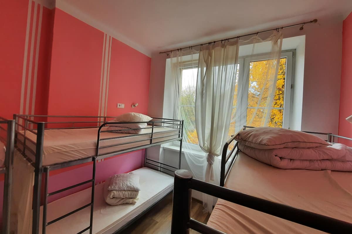 A dormitory room with bunk beds, a window with curtains, and a view of autumn foliage.