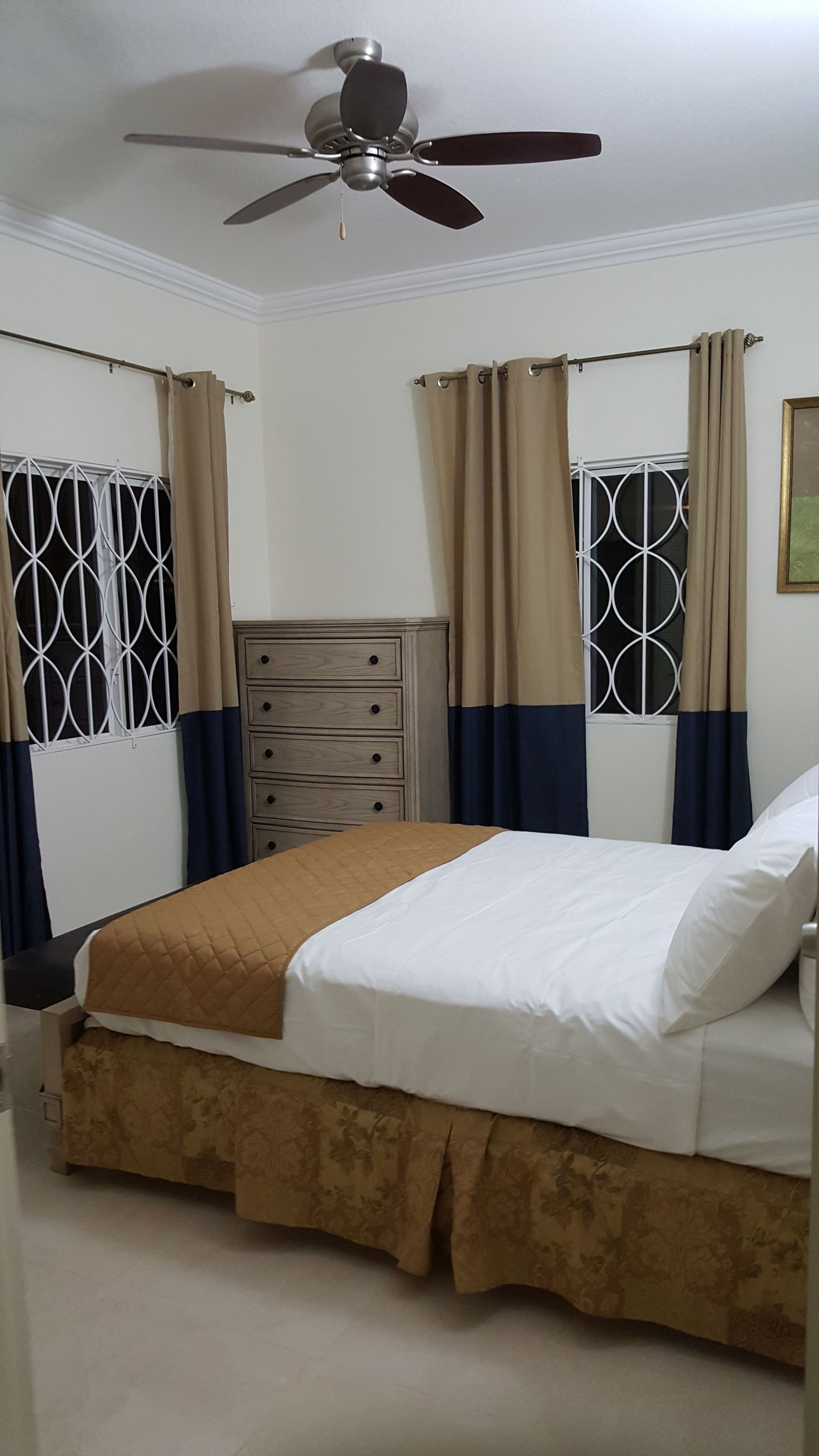Un dormitorio con cama, cómoda, ventilador de techo y ventanas con cortinas.