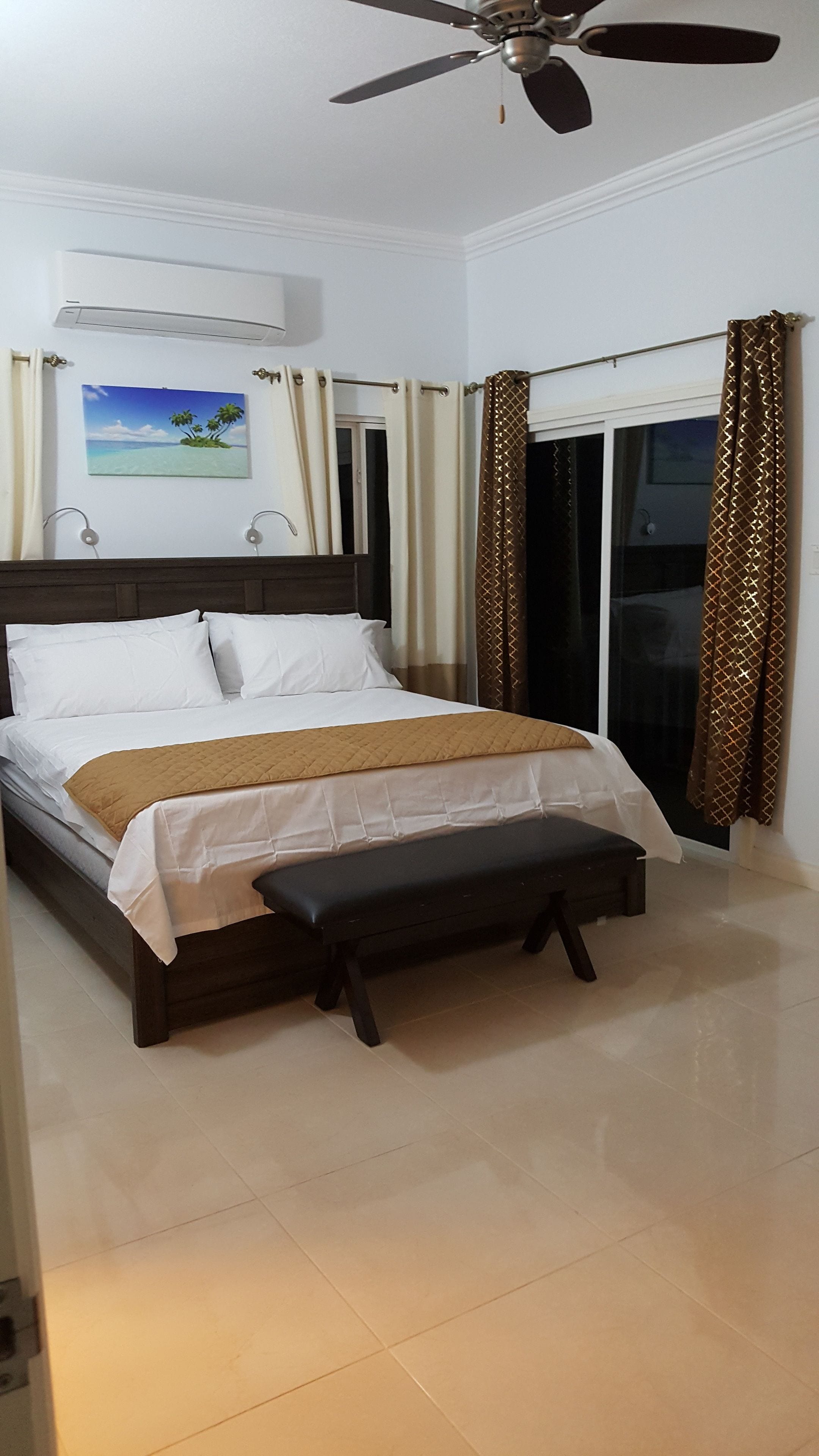 A bedroom with a bed, a ceiling fan, a window with curtains, and a painting on the wall.