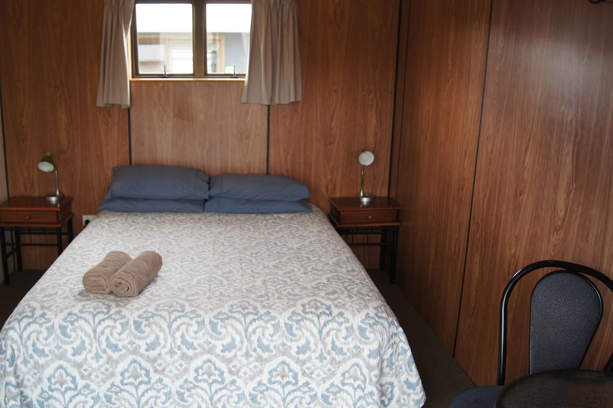 A bedroom with a wooden paneled wall, a bed with a patterned bedspread, two pillows, two bedside tables with lamps, and a chair.