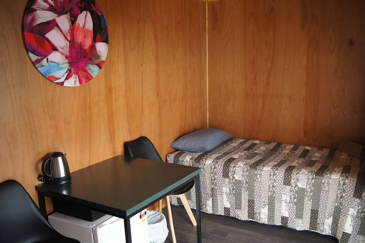 A small, compact room with a wooden wall, a bed with a patterned blanket, a desk with a kettle, and a chair.
