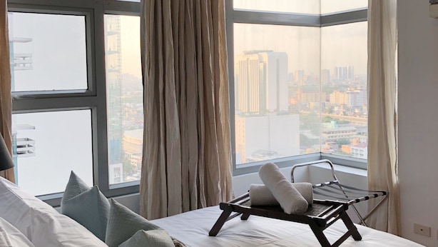 A hotel room with a large bed, a chair, and a view of a cityscape through large windows. - Cozy Pad in Heart of Makati HiSpeed WiFi (Makati)