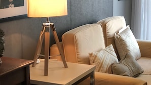 A modern living room with a beige sofa, a wooden side table with a lamp, and framed pictures on the wall. - Cozy Pad in Heart of Makati HiSpeed WiFi (Makati)