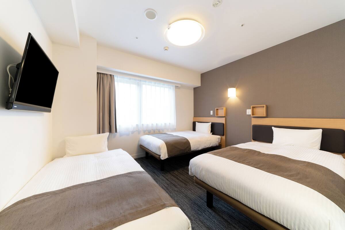 A hotel room with three beds, a flat-screen TV, a window with curtains, and a wall-mounted light fixture.
