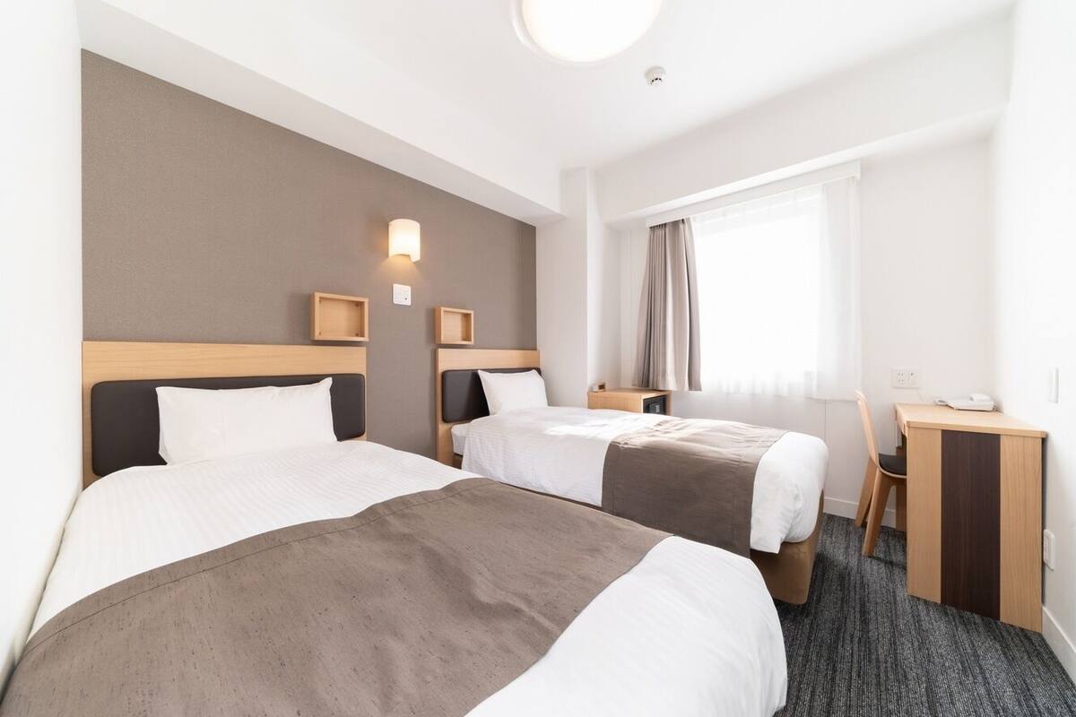 A hotel room with two beds, a desk, a chair, and a window with curtains.