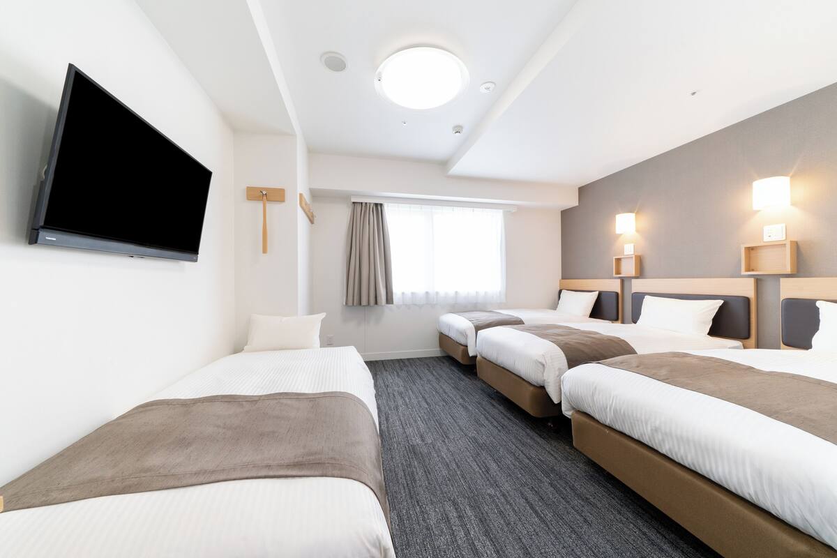 A hotel room with three beds, a flat-screen TV, a window with curtains, and wall-mounted lights.