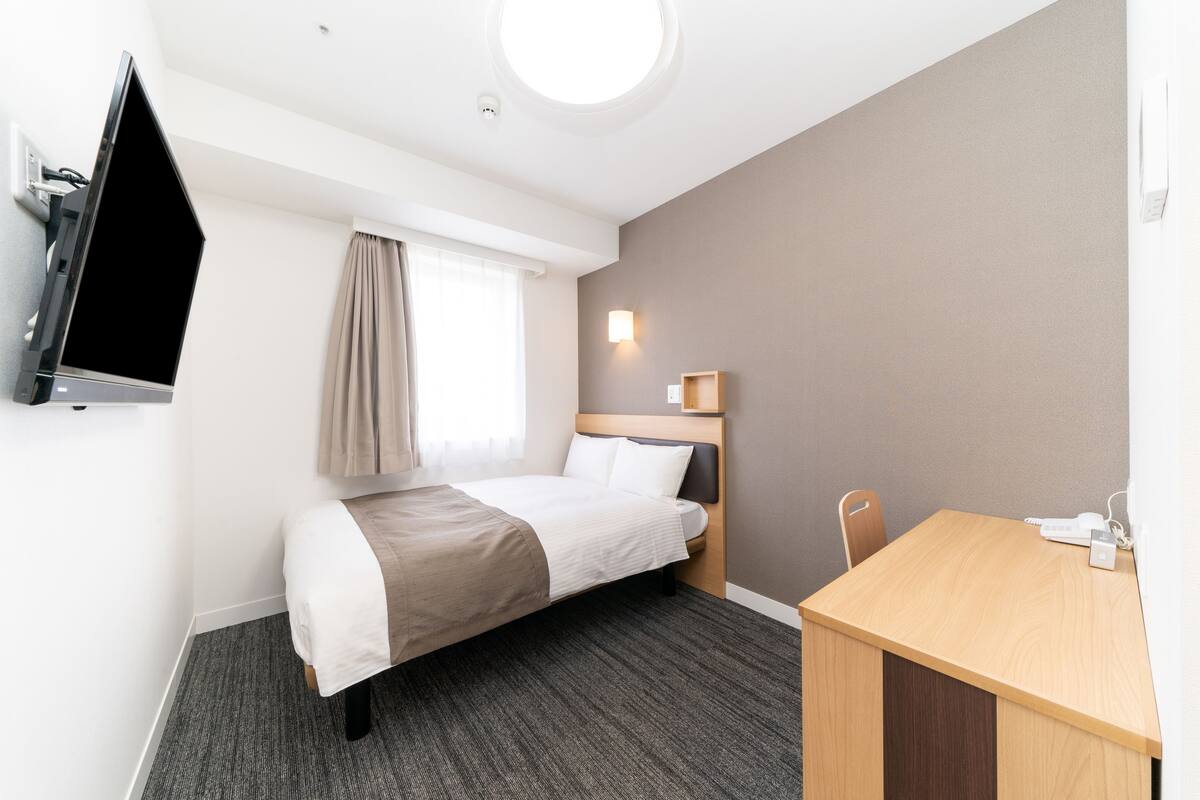 A hotel room with a bed, a desk, a flat-screen TV, a wall-mounted light, and a window with curtains.