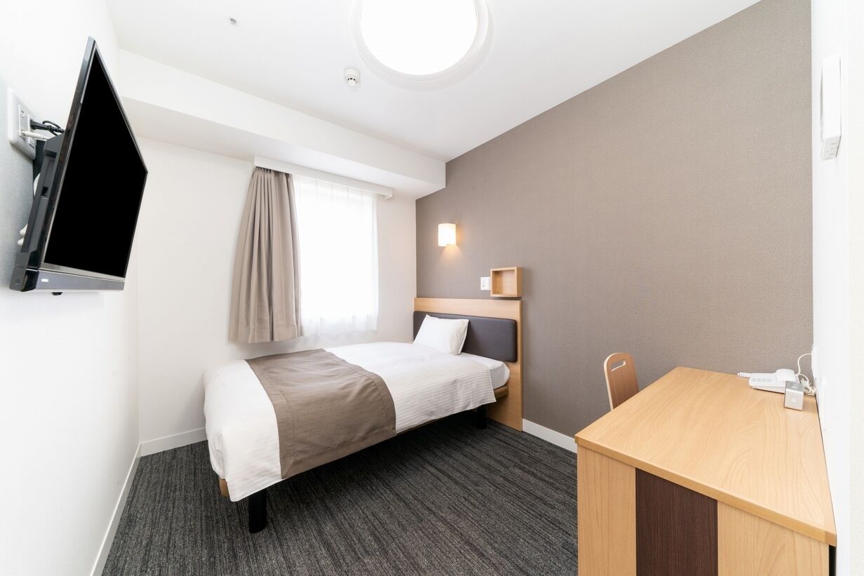 A hotel room with a bed, a desk, a television, a window with curtains, and a wall-mounted light.