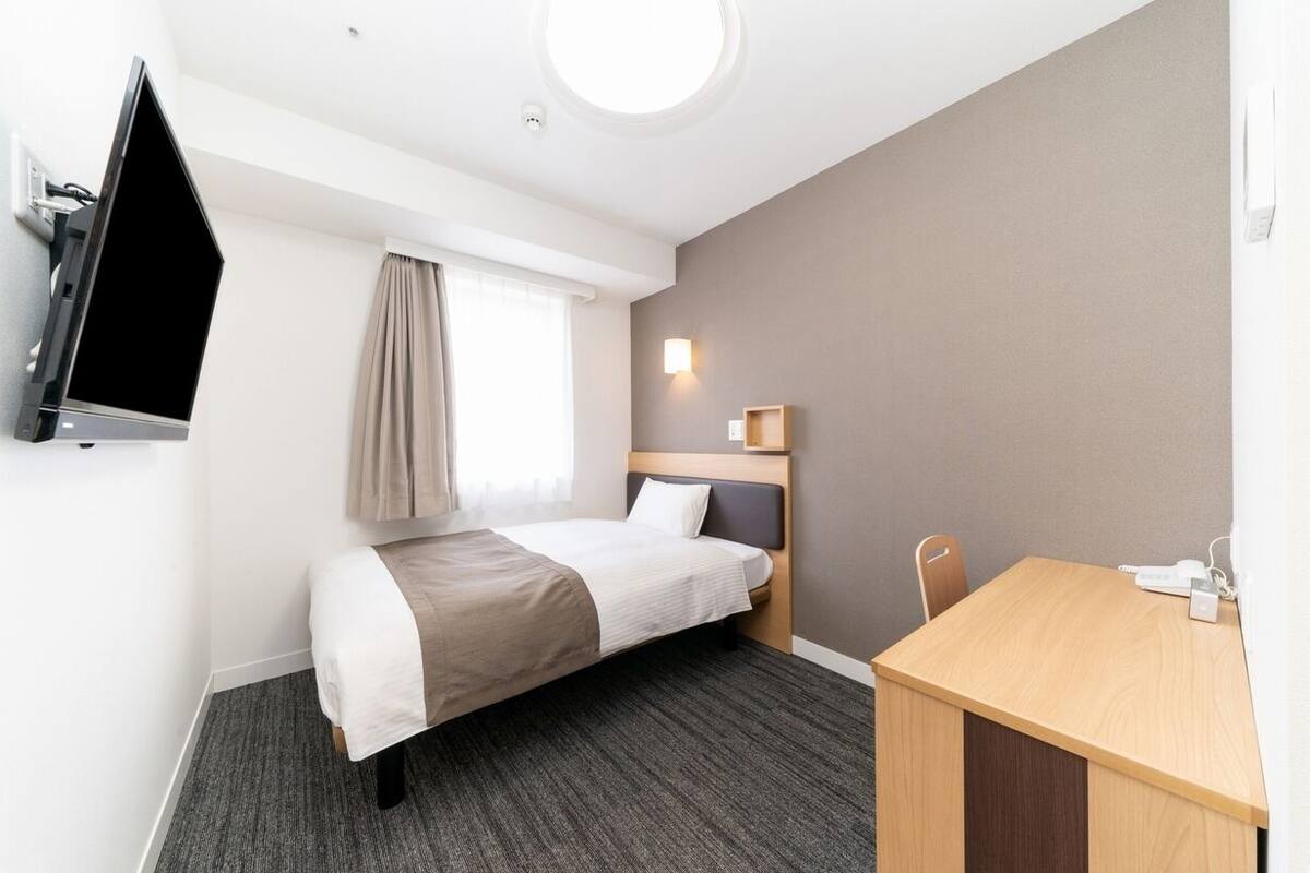 A hotel room with a bed, a desk, a television, a window with curtains, and a wall-mounted light.