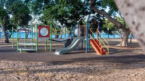 A playground with slides and climbing structures, surrounded by trees and a body of water.