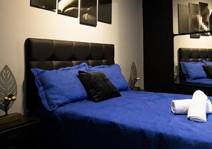 Una cama bien hecha con ropa de cama azul y una almohada negra, una toalla blanca y una mesita de noche con un objeto decorativo.