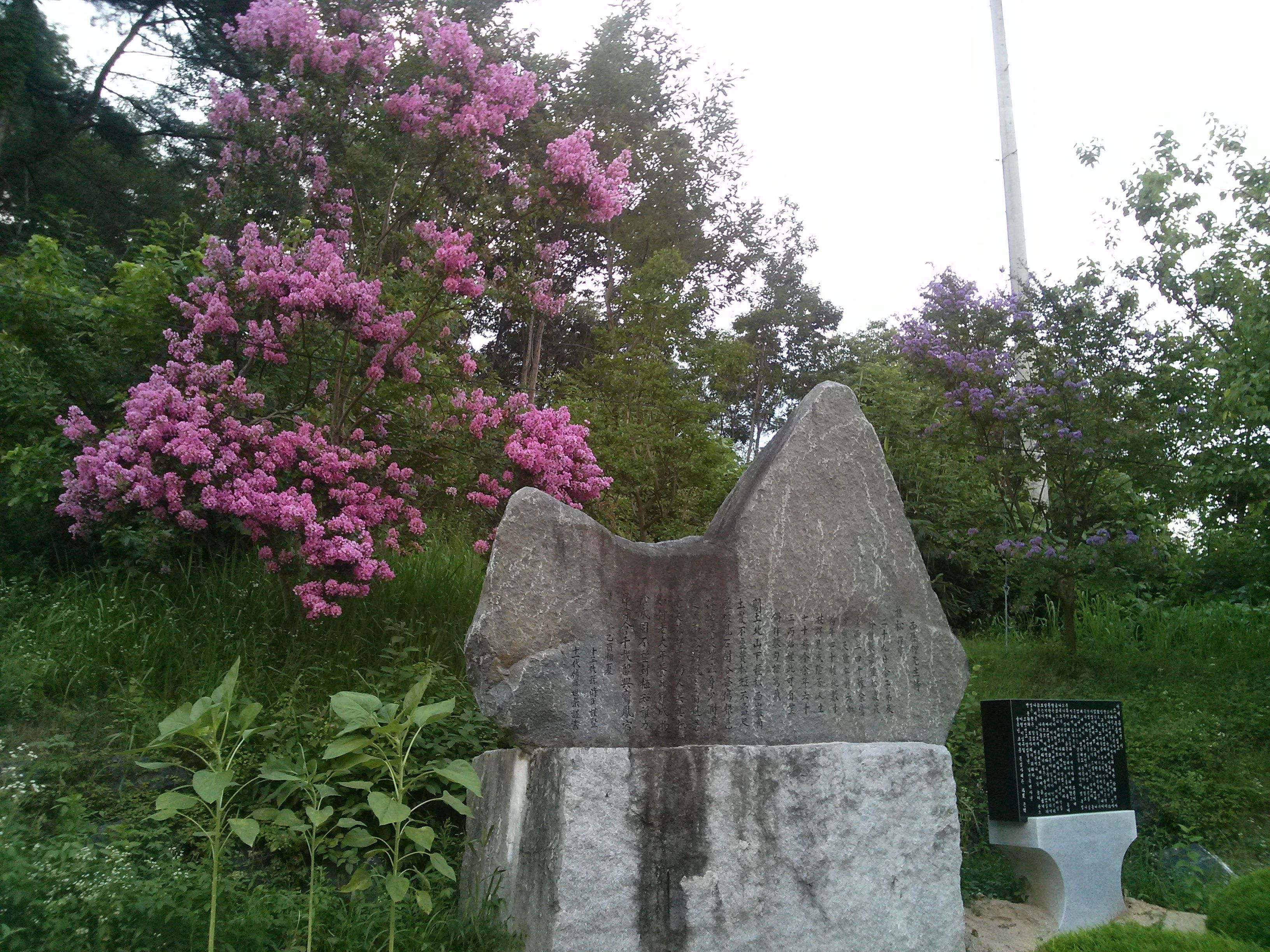 A stone monument with inscriptions, surrounded by lush greenery and flowering trees.