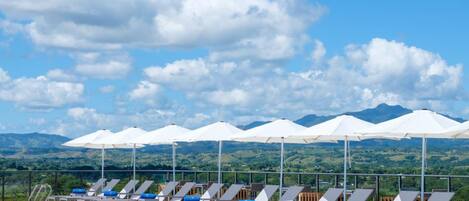A poolside area with lounge chairs and umbrellas, a clear sky, and a mountainous landscape in the background.