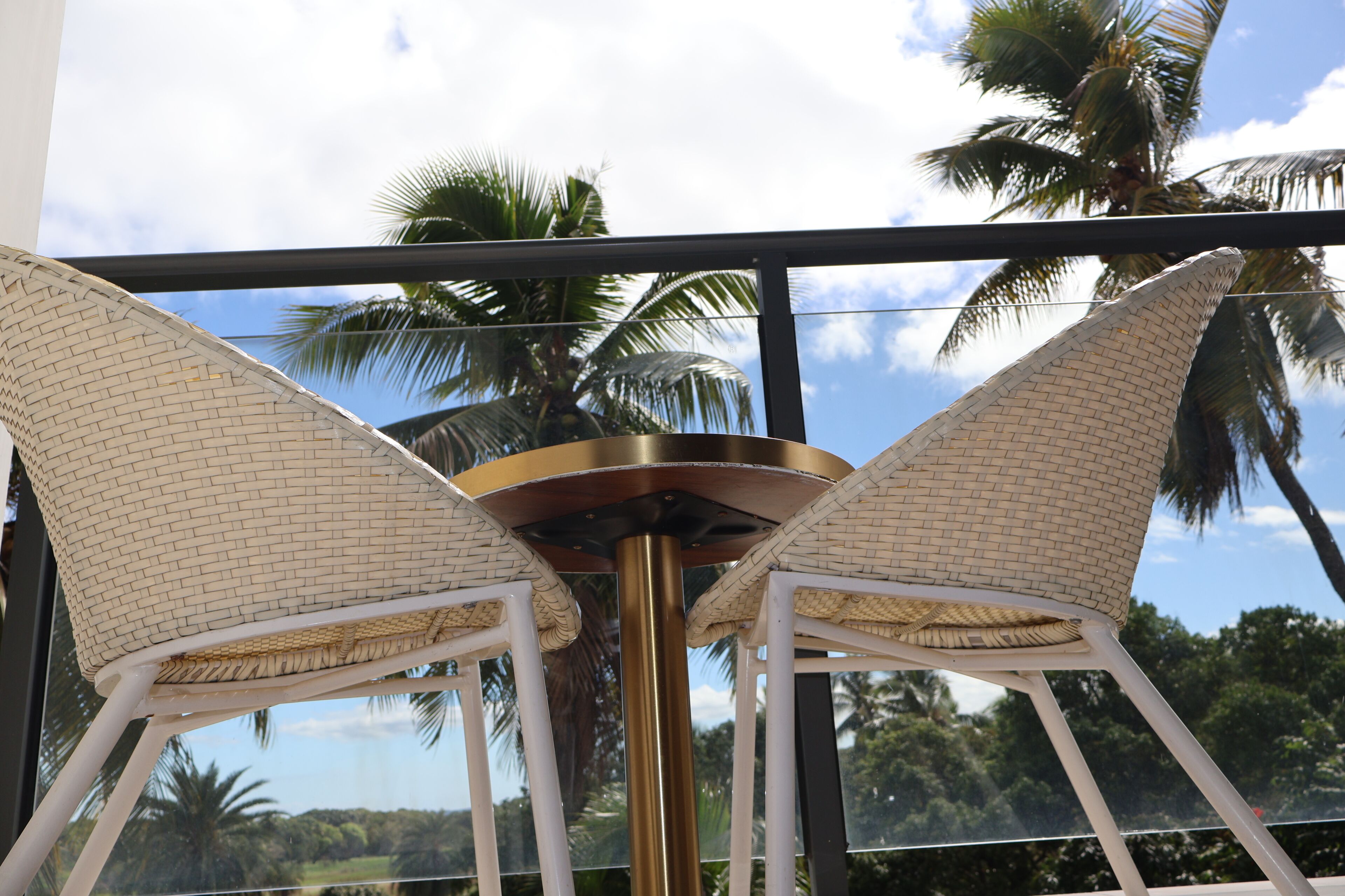 A view of a balcony with wicker chairs and a table, overlooking palm trees and a clear sky.