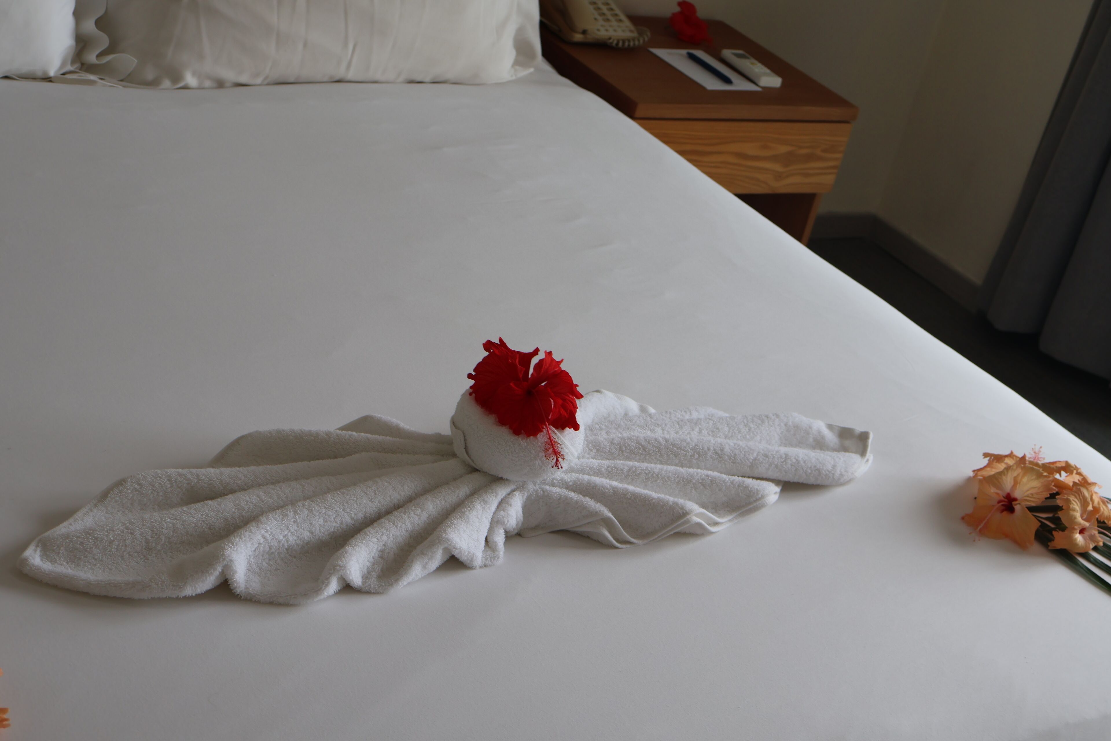 A neatly made bed with a towel arranged in a bow shape, a red flower, and a small orange flower on the side.