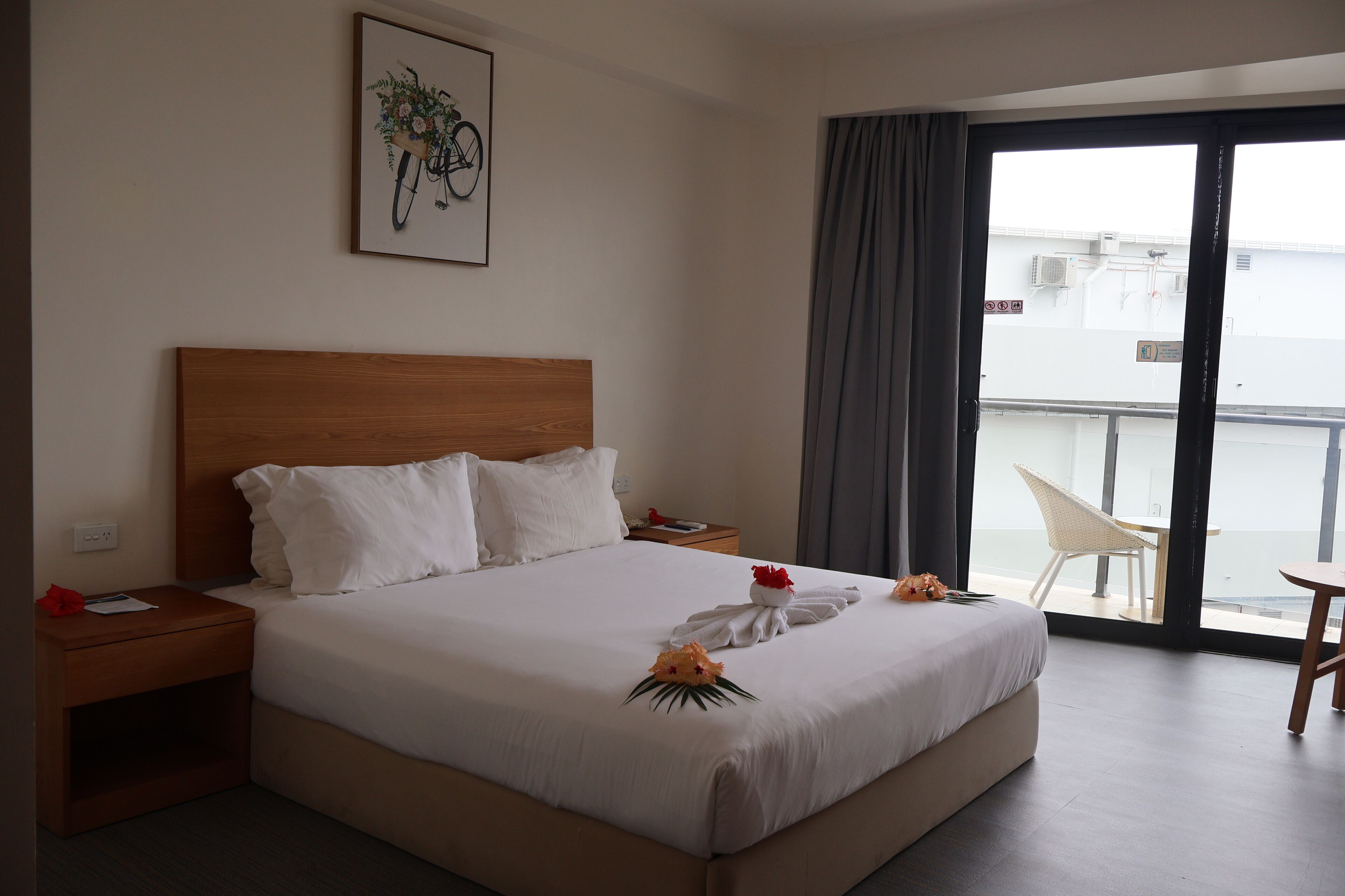 A hotel room with a large bed, a wooden headboard, white bedding, a framed picture on the wall, a nightstand with a red object, and a view of the outside through a sliding door.